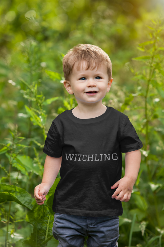 Toddler wearing black "Witchling" short sleeve tee outdoors.