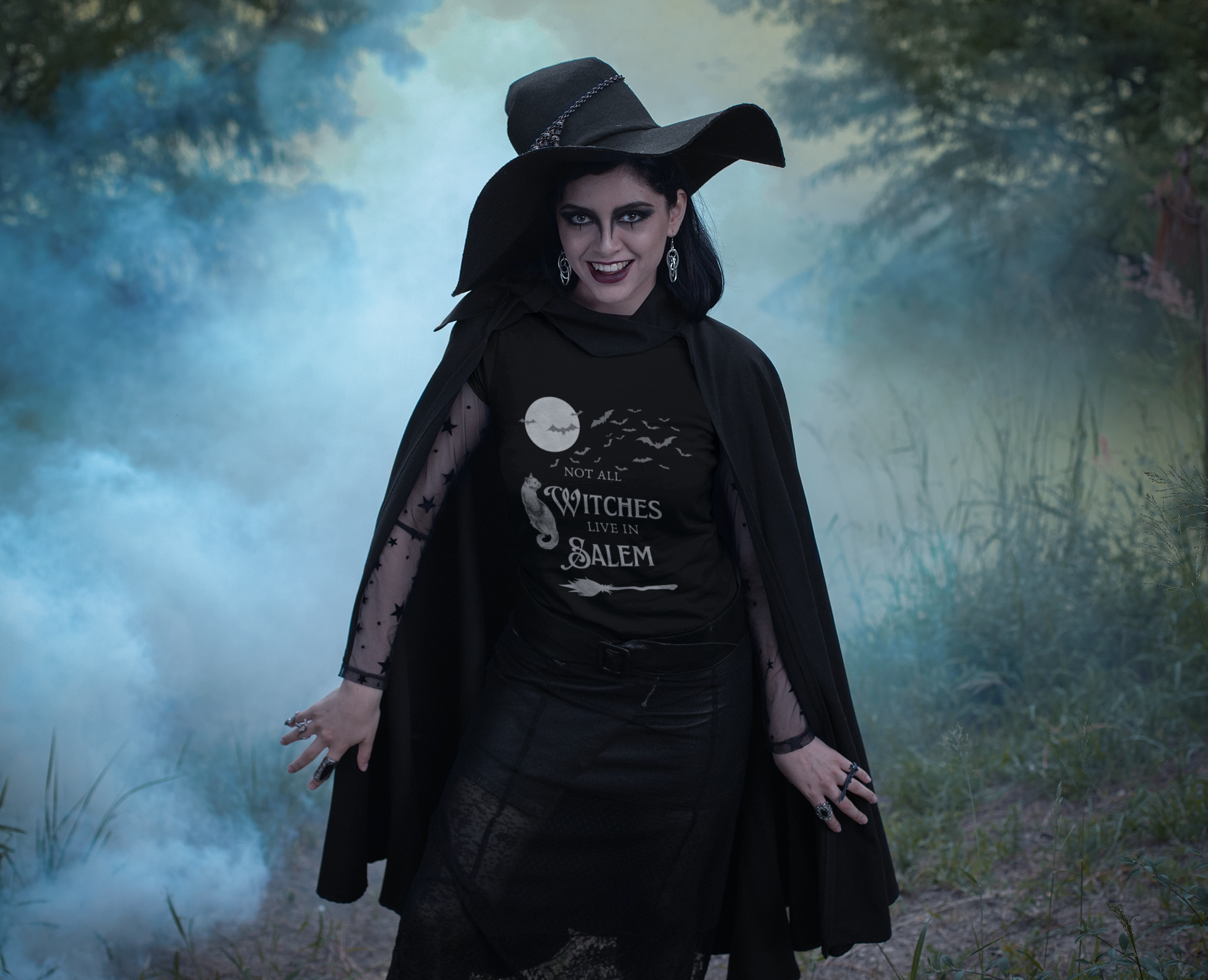 Woman wearing a black "Not All Witches Live in Salem" T-shirt with witch costume and cape in a foggy forest setting