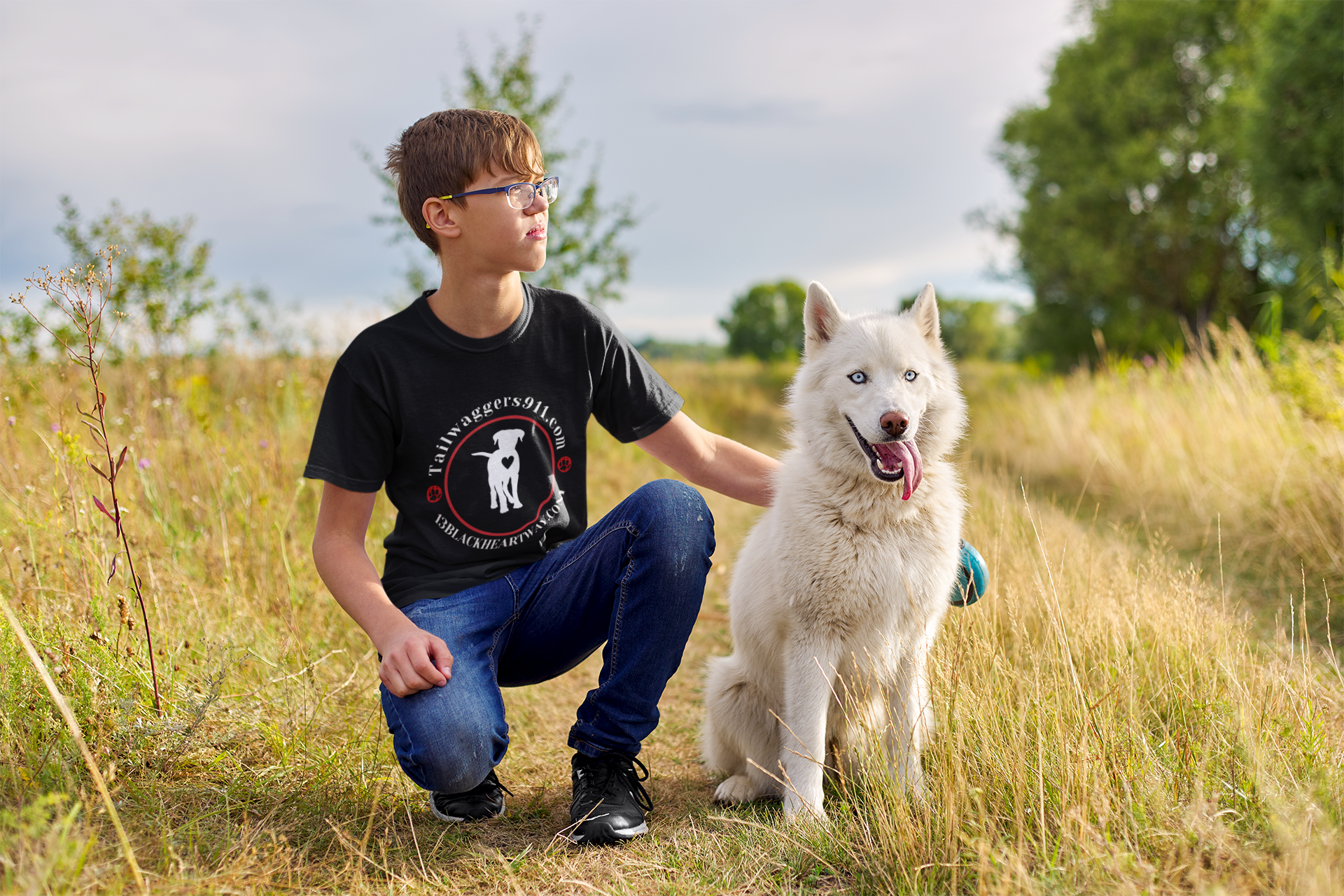 Child wearing black Tailwaggers 911 Kids Heavy Cotton Tee kneeling next to white dog in grassy outdoor setting.