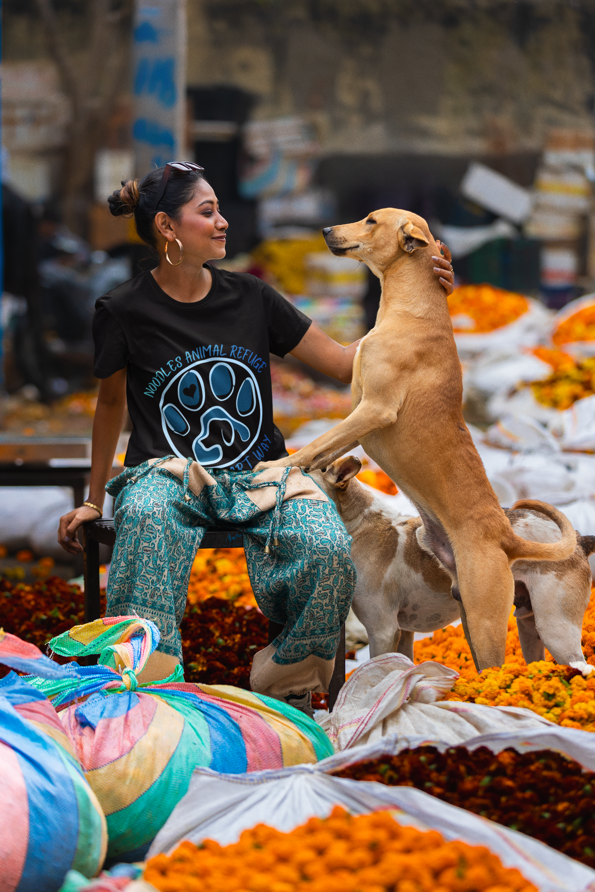 Woman wearing Look Good Do Good Tee with Noodles Animal Refuge logo, interacting with dogs at outdoor market with colorful flowers.