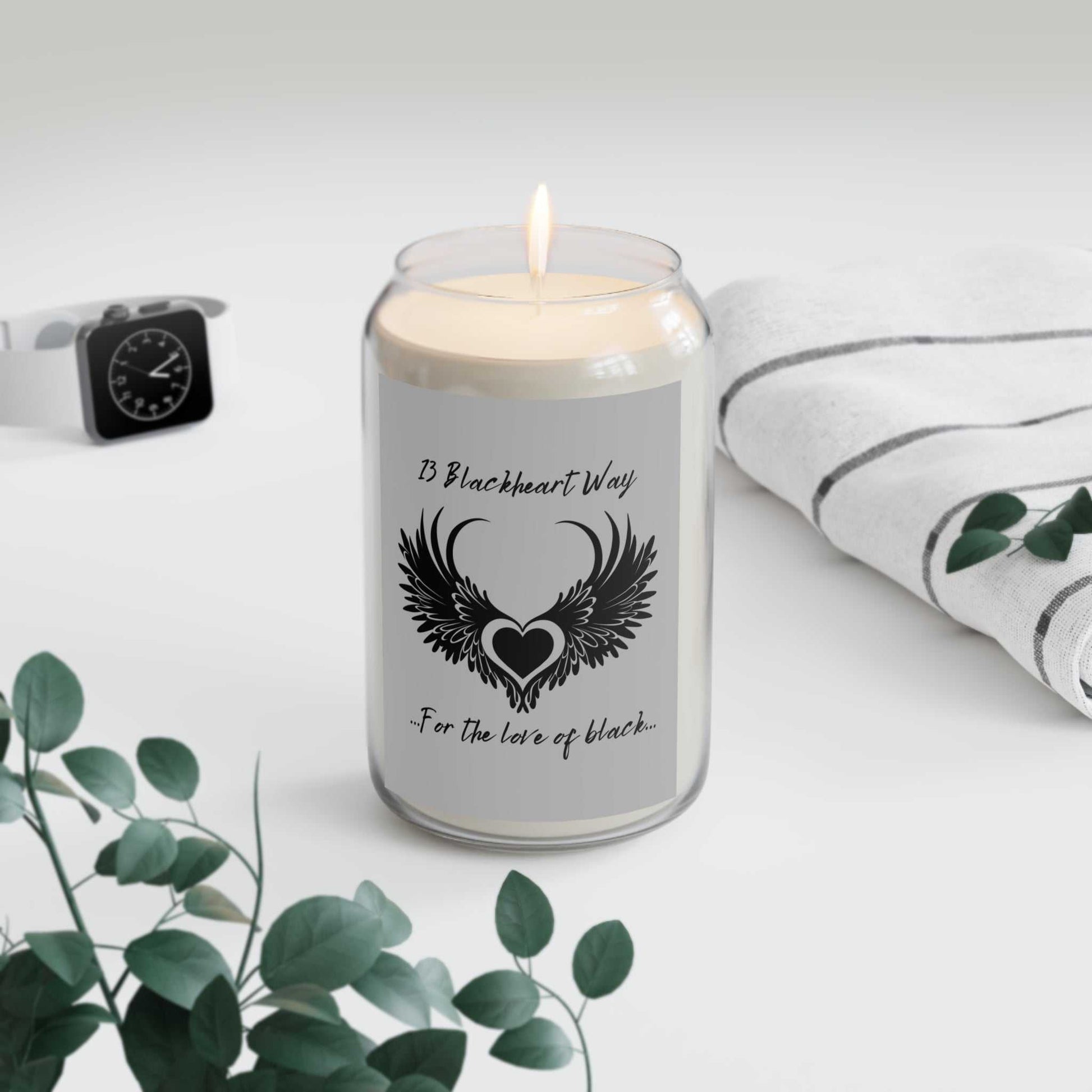 Blackheart Way Scented Candle in a glass vessel, featuring a heart with wings design, next to a striped cloth and greenery.